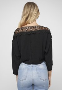 Svart topp med blonder langs skuldrene, lange ermer og en ledig passform, kombinert med lyseblå jeans med klassisk lommestil.