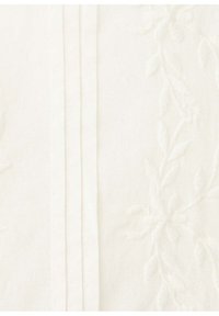 Tissu blanc avec des plis verticaux à gauche et une broderie florale subtile à droite montrant des feuilles et des fleurs.