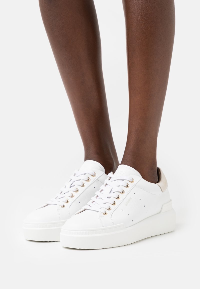 Baskets blanches à plateforme avec œillets dorés portées sur des jambes à la peau foncée, présentées sur un fond clair uni.