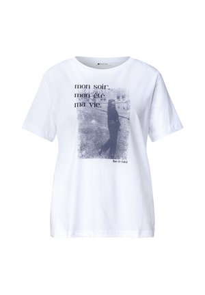 Femme blonde souriante regardant de côté, portant un t-shirt blanc uni à manches courtes et un jean bleu clair sur un fond uni.