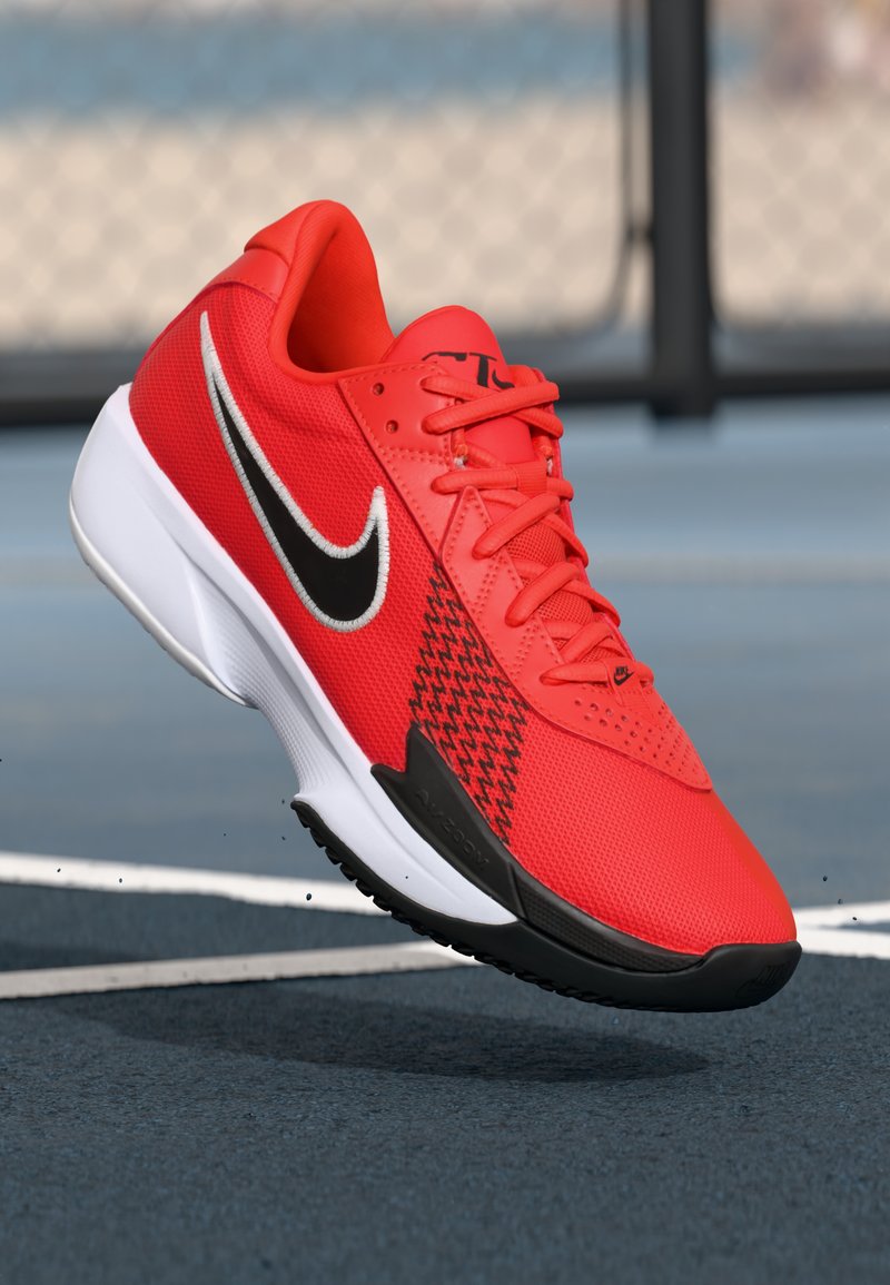 Rød atletisk sko med overdel i netmateriale, sort Nike swoosh, houndstooth-mønster på siden og hvid sål. Har teksturerede snørebånd og perforeringer.