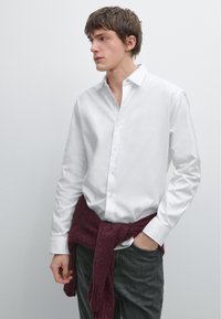 White button-up shirt with long sleeves, featuring a relaxed fit. A burgundy sweater tied around the waist adds texture and color contrast.