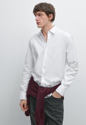 Camicia bianca button-up con maniche lunghe, caratterizzata da una vestibilità rilassata. Un maglione bordeaux legato in vita aggiunge texture e contrasto di colore.