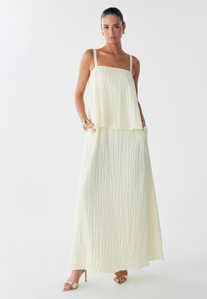Model in a flowing, light yellow maxi dress with textured fabric and thin straps, posed against a white background, wearing gold accessories.