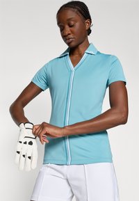 Světlemodrá polos tričko s krátkým rukávem a véčkovým výstřihem, se kontrastním pruhem na přední straně. Model si upravuje bílé golfové rukavice.