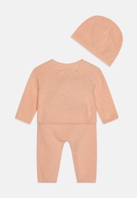 Nertinis dviejų dalių kostiumas švelnios persikų spalvos, su ilgomis rankovėmis, tekstūruota medžiaga ir derančia kepure. Apvalus iškirpimas ir raštuoti rankogaliai.