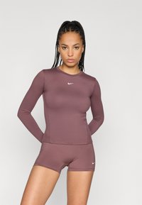 Dlhý rukáv športový top a šortky v tlmenom fialovom odtieni. Hladká tkanina s priliehavým strihom a bielym logom Nike na hrudníku a šortkách.