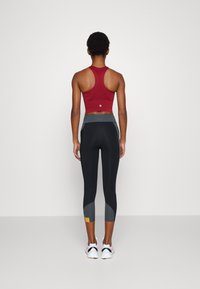 Bordowy sportowy crop top z teksturowanym wzorem i stylem racerback, zestawiony z czarnymi legginsami z szarymi akcentami i małą żółtą metką.