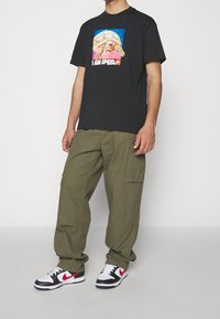 Camiseta de algodón negra con un diseño gráfico colorido que presenta una tortuga y el texto "SOY VELOCIDAD". Pantalones cargo verde oliva y zapatillas en blanco y negro.