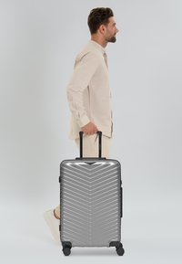 Silberner Hartschalenkoffer mit Zickzackmuster, ausziehbarem Griff, vier Rädern und seitlichem Griff, gehalten von einer Person in einem hellen Hemd.