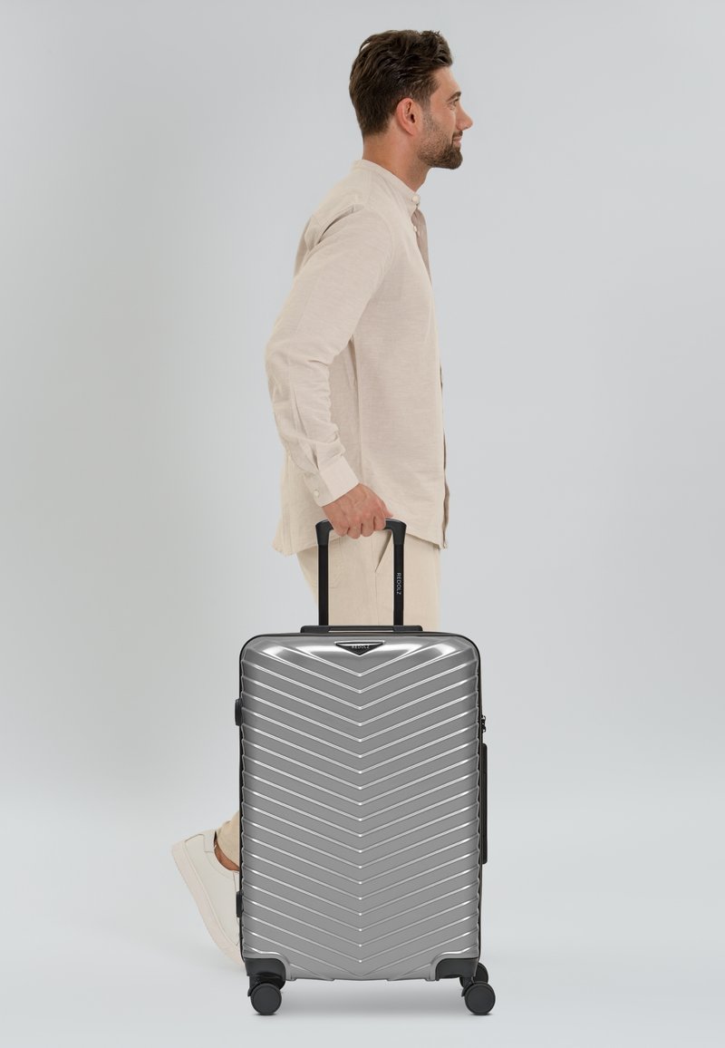 Silberner Hartschalenkoffer mit Zickzackmuster, ausziehbarem Griff, vier Rädern und seitlichem Griff, gehalten von einer Person in einem hellen Hemd.