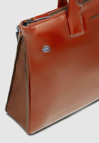 Braune Ledertasche mit strukturiertem Design, ausgestattet mit einem Reißverschluss, kontrastierenden blauen Akzenten und einem Logodetail auf der Vorderseite.