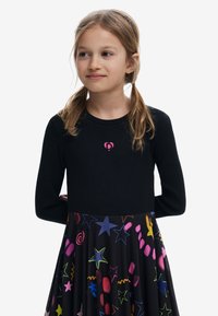 Ung pige med lyst brunt hår i to fletninger iført en sort bluse med lange ærmer med en pink hjerte-logo og et farverigt nederdel med stjerneprint.