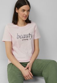 T-shirt rose clair à manches courtes avec du texte en sequins noirs "beauty of being". Le t-shirt a un col rond et est associé à un pantalon vert.