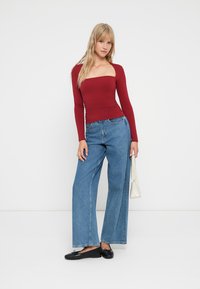 Maglietta rossa a maniche lunghe con scollatura quadrata, abbinata a pantaloni in denim blu a gamba larga. Modella che tiene una piccola borsa bianca, indossando ballerine nere.