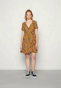 Vestido envolvente de manga curta em castanho com padrão de leopardo em preto e creme, apresentando um laço na cintura e uma saia rodada. Combinado com ténis pretos.