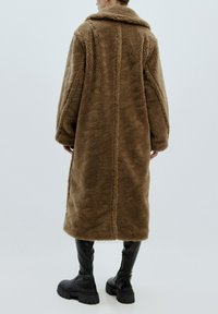 Brauner Kunstpelzmantel mit breitem Kragen, langen Ärmeln und lockerem Schnitt. Auffällige Nähte schaffen ein strukturiertes Muster entlang des Rückens.