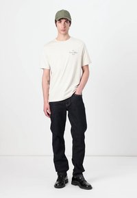 Tricou crem cu un model texturat deschis, blugi închis, și o șapcă verde. Pantofi negri lustruiți, stând pe un fundal alb simplu.