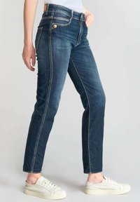 Jean en denim bleu foncé avec une coupe slim, comportant des poches avant, des finitions en or et des coutures contrastantes, associés à des baskets blanches.