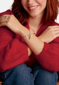 Silbernes Charm-Armband mit verschiedenen Anhängern, darunter eine Perle und dekorative Akzente, getragen über einem roten Wollpullover und Jeans.