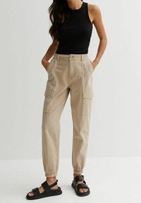Beige cargo broek met elastische boorden, twee zijzakken en een knoopsluiting aan de voorkant, gecombineerd met een zwart mouwloos topje en zwarte sandalen.