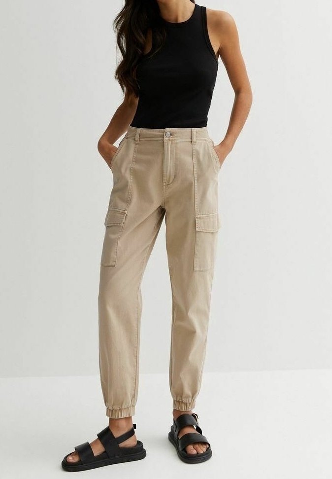 Beige cargo broek met elastische boorden, twee zijzakken en een knoopsluiting aan de voorkant, gecombineerd met een zwart mouwloos topje en zwarte sandalen.