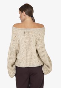 Donna con capelli castani raccolti, che indossa un maglione chunky a maniche scoperte di colore beige e pantaloni scuri, rivolta di spalle su uno sfondo bianco.