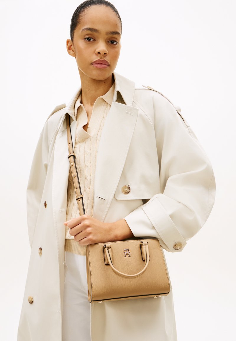 Γυναίκα που φοράει μπεζ καμπαρντίνα και πουλόβερ με πλέξη καλώδιου κρατά μια ταμπά δερμάτινη τσάντα με ιμάντα ώμου μπροστά σε λευκό φόντο.