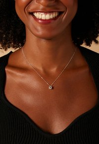 Zilveren ketting met een vierkant hangertje met een centrale steen, gedragen op een textuurrijke zwarte top, waarbij de huidtinten en halslijn worden benadrukt.