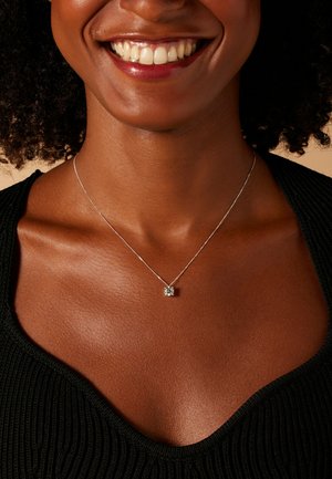 Zilveren ketting met een vierkant hangertje met een centrale steen, gedragen op een textuurrijke zwarte top, waarbij de huidtinten en halslijn worden benadrukt.