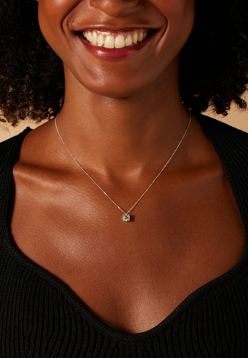 Zilveren ketting met een vierkant hangertje met een centrale steen, gedragen op een textuurrijke zwarte top, waarbij de huidtinten en halslijn worden benadrukt.