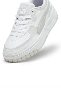Baltas sneakers ar teksturētu ādu un acu akcentiem. Iespējas ietver biezu gumijas zoli, šņorēšanas dizainu un diskrētu logotipa detalizāciju.