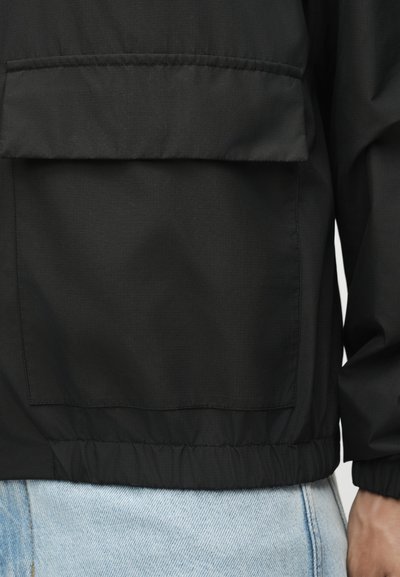 Μαύρο μπουφάν με μεγάλη μπροστινή τσέπη και ελαστική μέση, φορεμένο πάνω από ανοιχτό μπλε τζιν, το χέρι είναι ορατό στο πλάι.