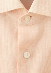 Svetlo broskyňová textúrovaná látka s detailom goliera košele a dvoma bielymi gombíkmi zapnutými zvisle na prednej légii.