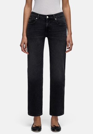 Kobieta ubrana w czarne jeansy o prostych nogawkach, biały włożony do środka top i czarne baleriny, stoi na tle jednolitego tła.