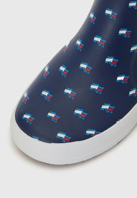 Chaussure bleu marine avec une finition lisse et brillante, ornée d'un motif répété de petites drapeaux stylisés en rouge, blanc et bleu. Semelle blanche.