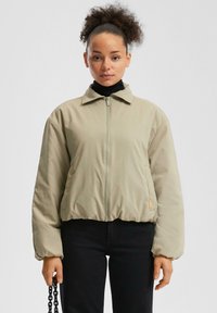 Veste courte beige clair avec une fermeture éclair à l'avant, un col pointu et une taille élastique. Tissu lisse et mat avec une coupe décontractée et des manches longues.