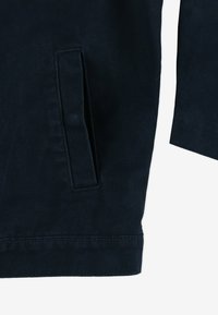 Granatowa kurtka z fakturowanej tkaniny, z kieszenią na suwak po lewej stronie i prostym rękawem bez widocznych elementów metalowych.