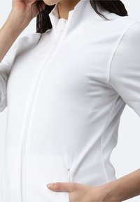 Giacca bianca con zip e colletto alto, caratterizzata da una texture liscia e un design aderente. Include tasche laterali e dettagli a costine.