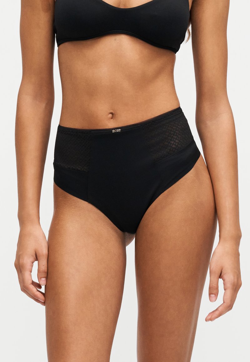 Femme portant un sous-vêtement noir taille haute avec des panneaux latéraux en mesh et un petit logo décoratif sur la ceinture avant.