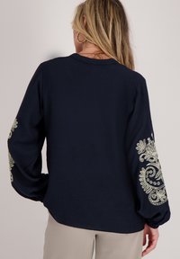 Marineblauwe blouse met lange mouwen, voorzien van ingewikkeld crèmekleurig borduurwerk op de manchetten, gemaakt van een lichtgewicht stof.