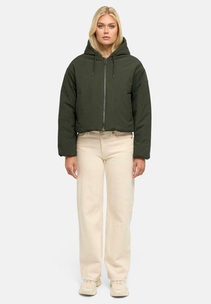 Femme debout portant une doudoune à capuche vert foncé, un pantalon droit taille haute beige et des baskets beige clair sur fond blanc.