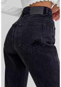 Black high-waisted jeans made of denim, featuring a distressed patch on the back pocket, visible stitching, and a logo embossed leather patch.