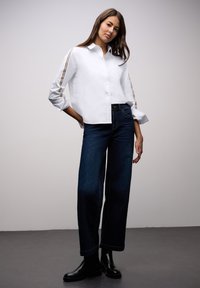 Vit skjorta med knappar och uppvikta ärmar, med bruna randiga detaljer. Kombinerad med vida mörkblå jeans och svarta stövlar.