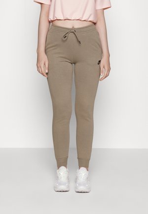 Woman wearing beige Nike joggers with drawstring, cropped pink top, and white sneakers, standing against a plain white background.