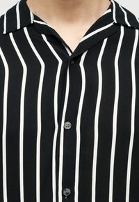 Κοντινή εικόνα μαύρου πουκάμισου με κάθετες λευκές ρίγες και μαύρα κουμπιά, που δείχνει το γιακά και το πάνω μέρος του στήθους.