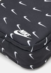 Svart Nike-väska med strukturtyg och upprepade vita swoosh-logotyper, rektangulär form och vadderad design för förvaring.