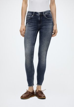 Femme portant un jean skinny bleu foncé avec un ourlet brut et des chaussures bateau en cuir marron sur un fond clair uni.