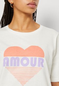 Vit t-shirt med kort ärm som har en stor korallfärgad hjärtgrafik med ordet "AMOUR" i lila, omgiven av tunna linjemönster nedanför.