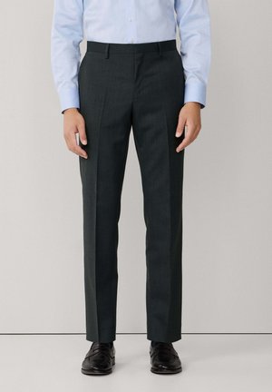 Pantalones rectos de color verde oscuro con una textura suave, que presentan un diseño de pierna recta, pliegues frontales y pasadores para cinturón, combinados con zapatos negros.